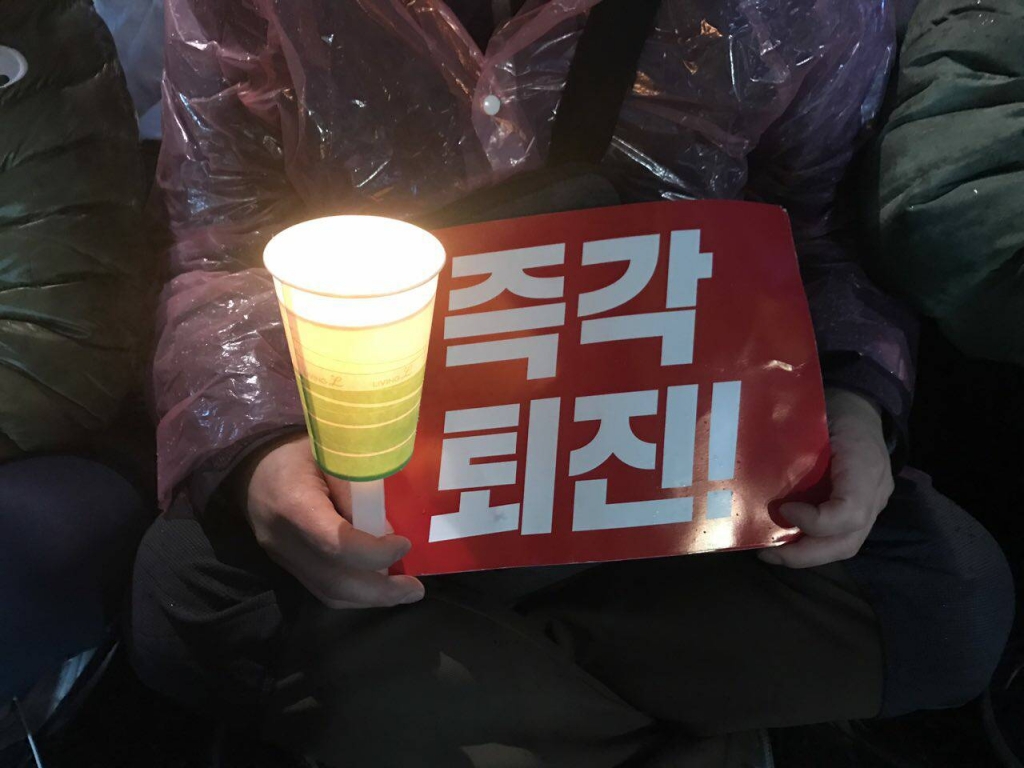 靜坐市民身穿輕便雨衣,一手拿著紙杯做的燭臺,一手緊掐"立馬下臺" (즉각 퇴진) 的紙板。圖/Lee Ji Eun